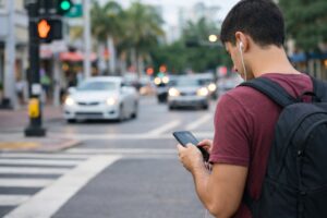 Pedestrian focused on a smartphone while standing at a city crosswalk with nearby traffic and signals visible.