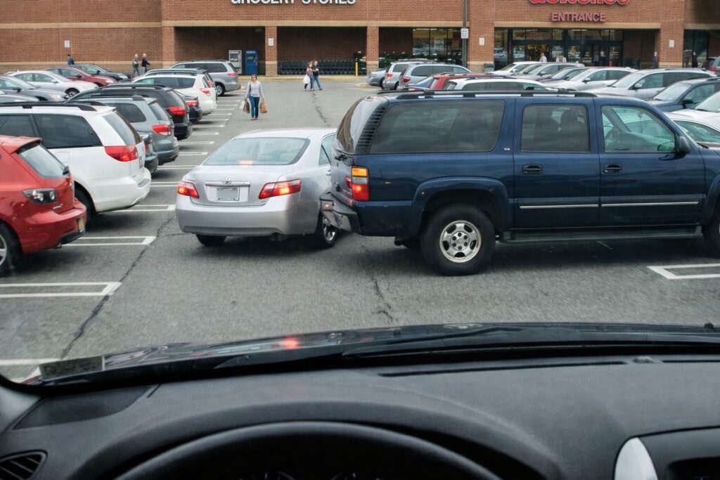 View from inside a car in a busy grocery store parking lot where a blue SUV has backed into a silver sedan while people walk toward the store entrance in the background.