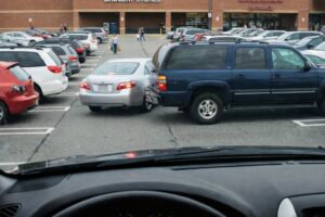 View from inside a car in a busy grocery store parking lot where a blue SUV has backed into a silver sedan while people walk toward the store entrance in the background.